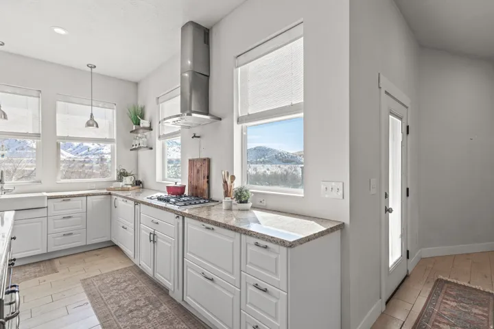 Contemporary kitchen featuring white cabinetry, light-toned countertops, a built-in gas range with an overhead vent hood, and multiple windows providing natural light