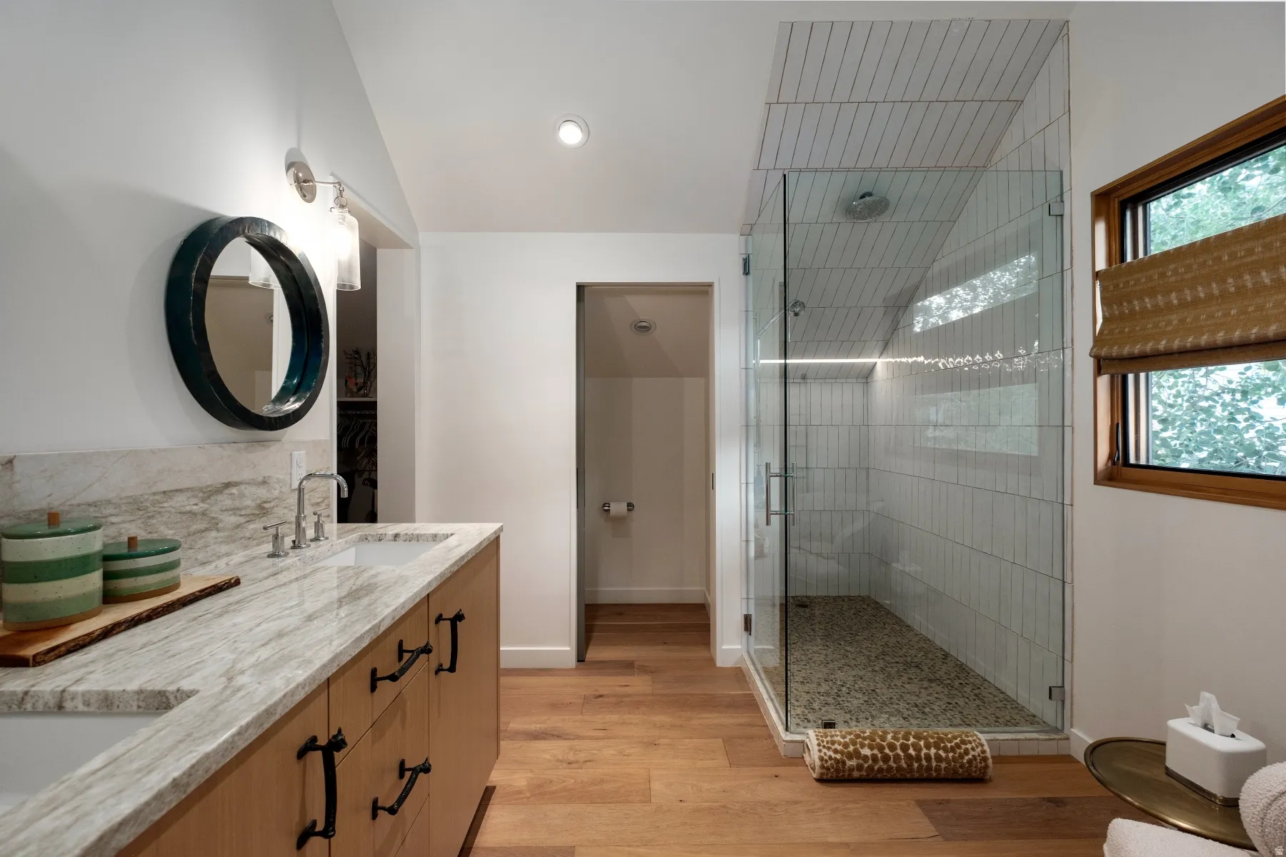 Spacious bathroom featuring a double vanity with stone countertop, wood-finish flooring, and a glass-enclosed shower with pebble flooring and tiled walls