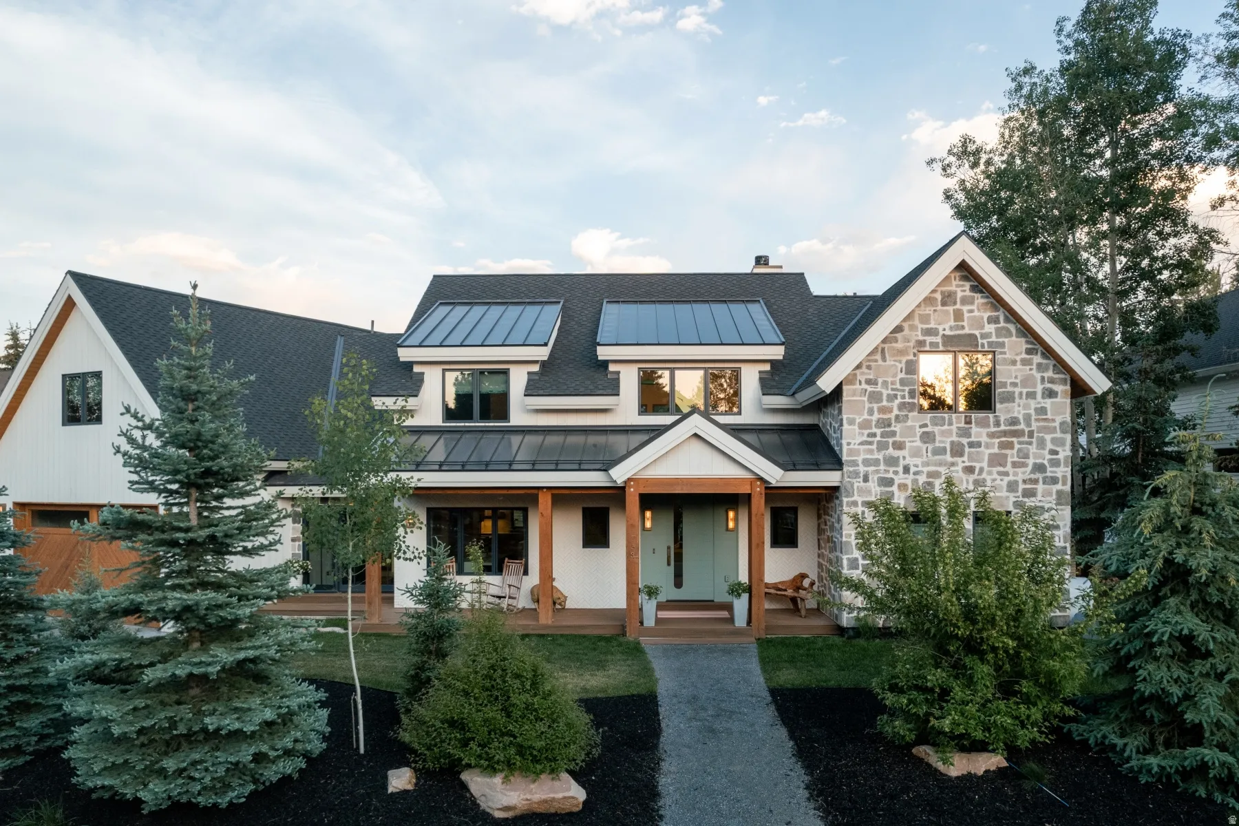 Modern farmhouse-style exterior featuring a stone facade, white vertical siding, dark shingled roof, metal roof accents, and an inviting covered entry porch with wood columns