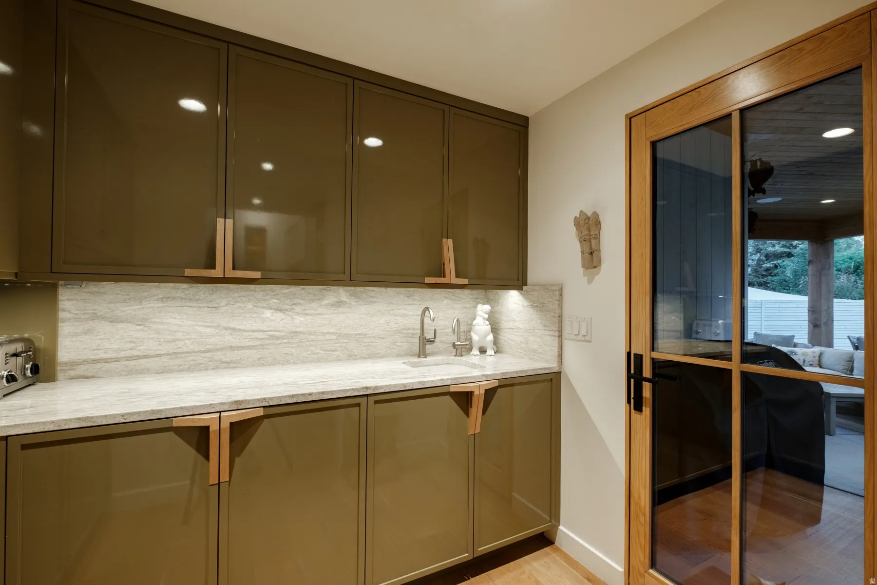 Modern wet bar featuring high-gloss olive cabinetry, integrated lighting, a stone countertop, a matching stone backsplash, and a stainless steel undermount sink with a gooseneck faucet