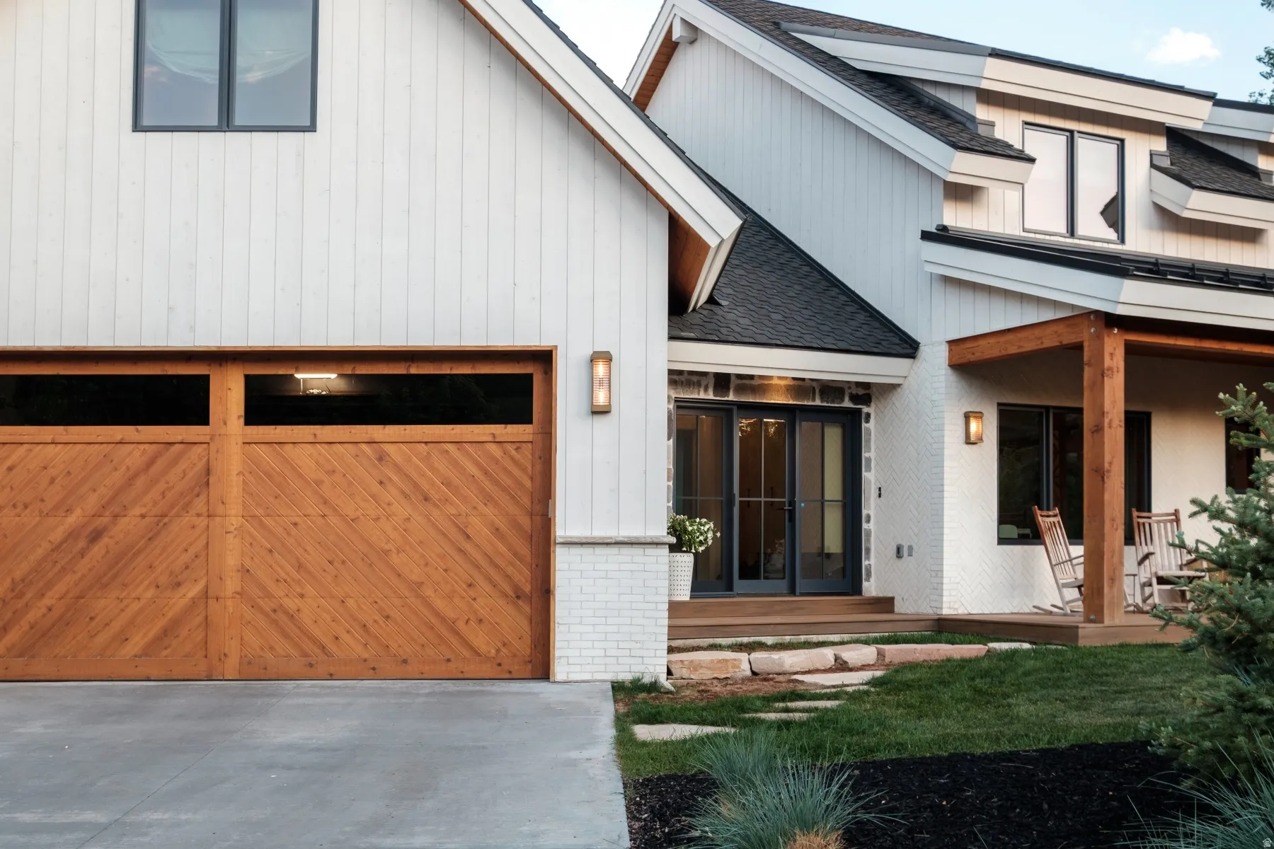 Contemporary farmhouse exterior featuring vertical white siding, a wood-finish garage door with upper window panels, and a covered porch with exposed wood beams