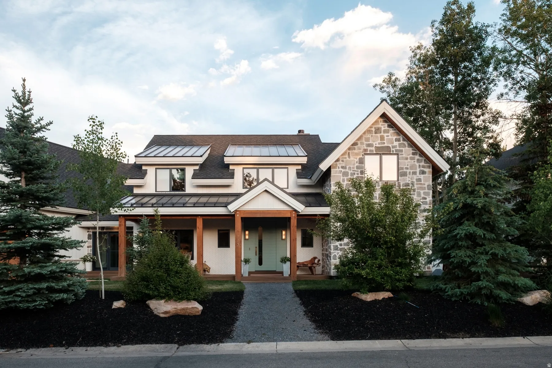 Craftsman-style exterior featuring a stone facade, dormer windows with metal accents, a covered front porch with natural wood columns, and a light-colored entry door