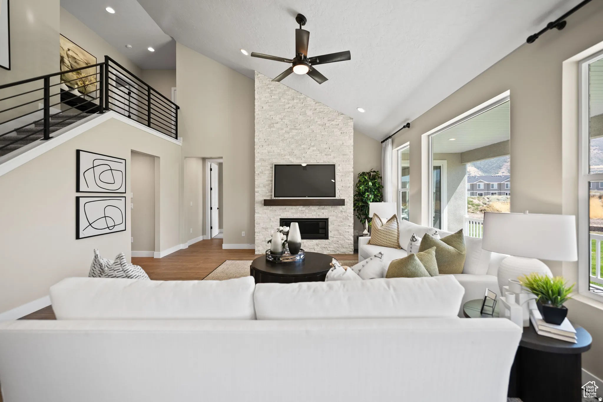 Vaulted ceiling living room featuring a stone-clad fireplace with wood mantel, large windows, a ceiling fan, and hardwood flooring