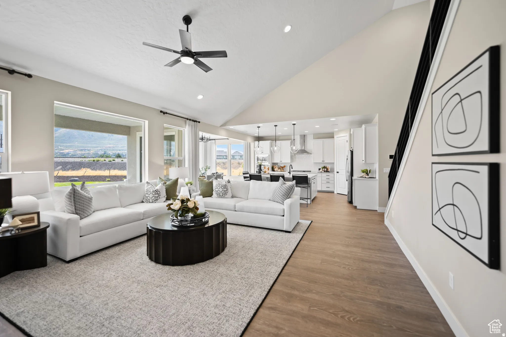 Spacious living area featuring a vaulted ceiling with a ceiling fan, large windows, wood-finish flooring, a contemporary staircase with black railings, and an open concept layout into the kitchen
