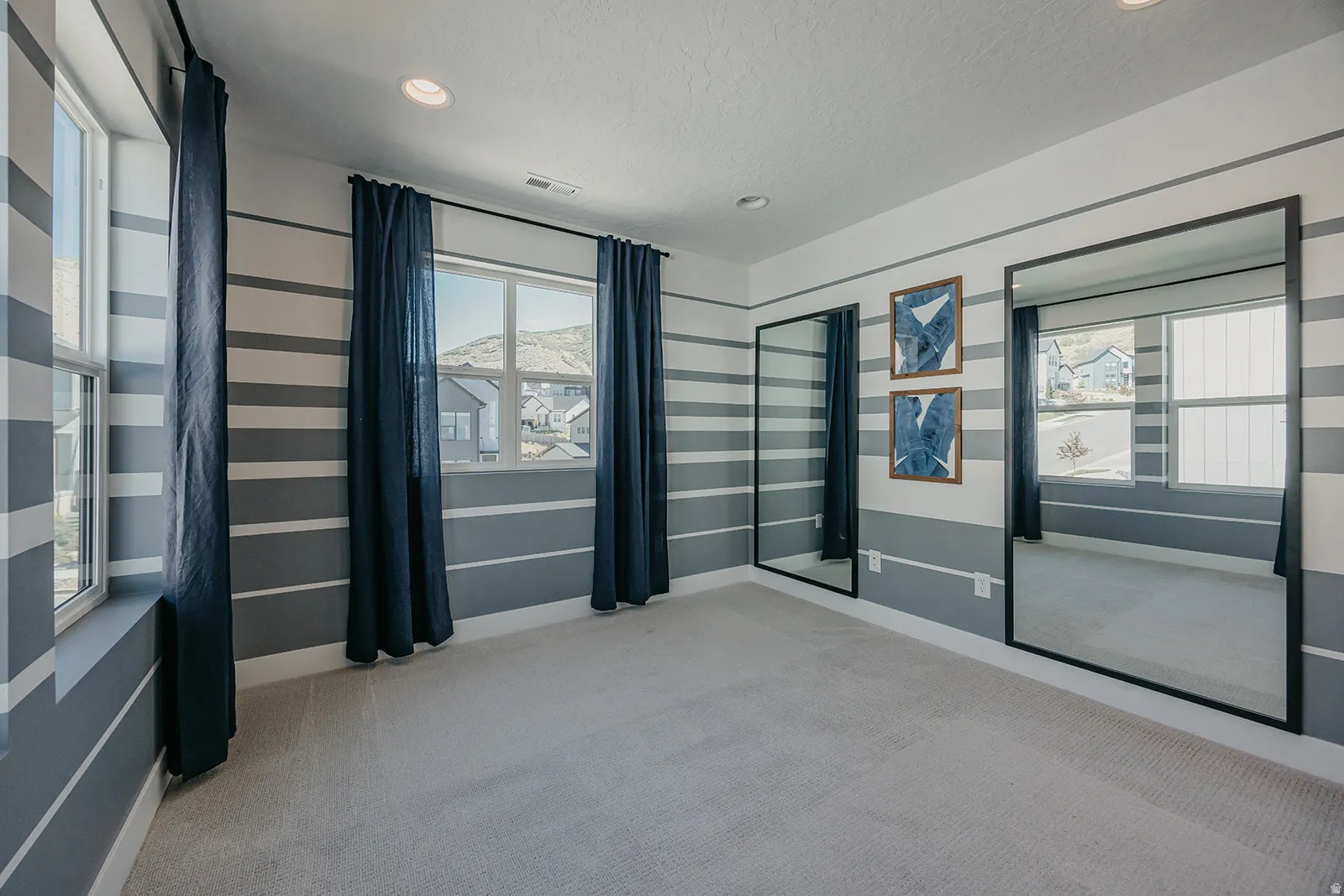Spacious room featuring light-colored carpeting, horizontal striped wall accents, and multiple windows providing natural illumination