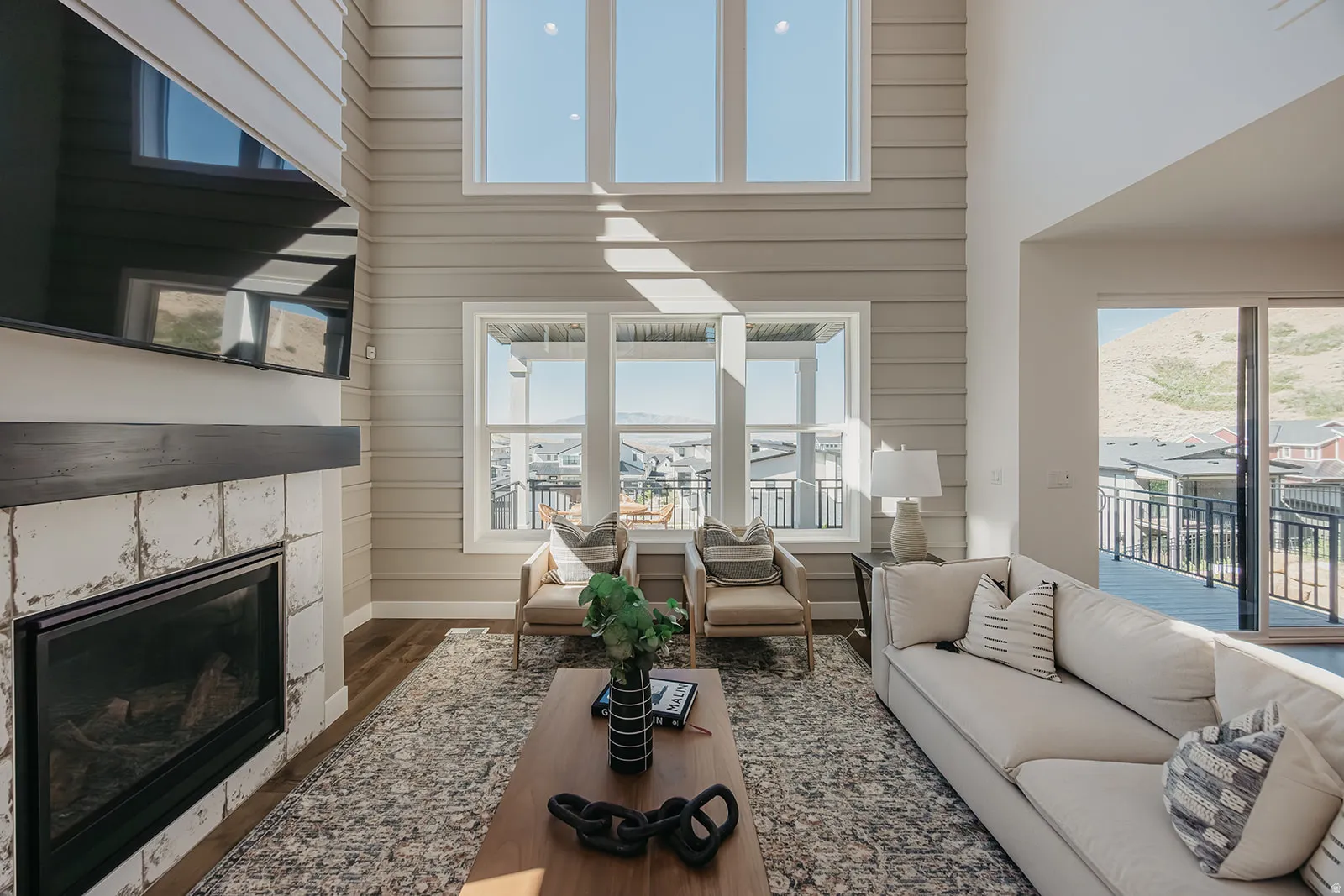 Two-story living area featuring a fireplace with white stone surround and dark wood mantel, wood-finish flooring, high ceilings with upper windows, and a sliding glass door opening to an outdoor deck