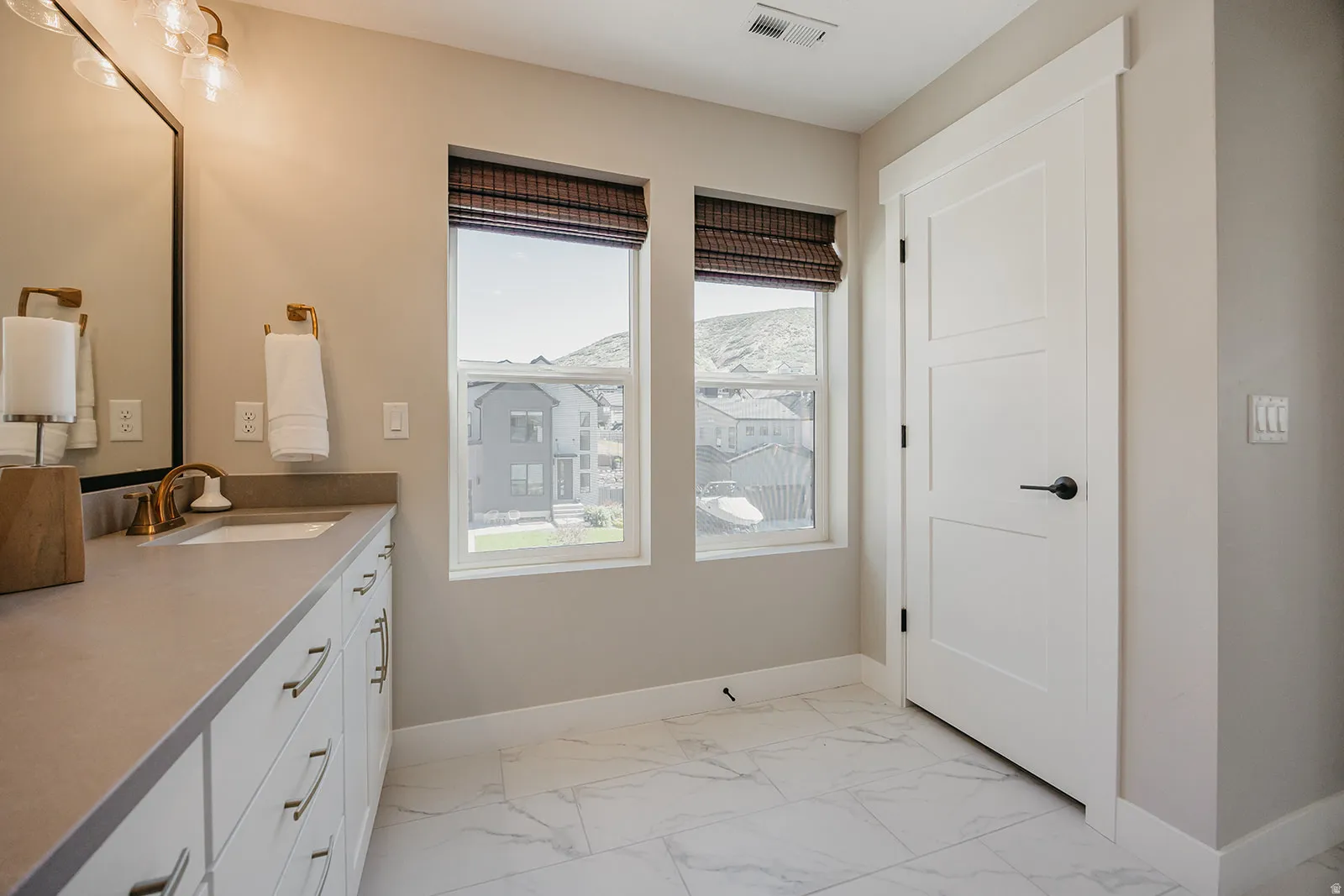 Contemporary bathroom featuring a single vanity with a gold-tone faucet, a large mirror, and two windows