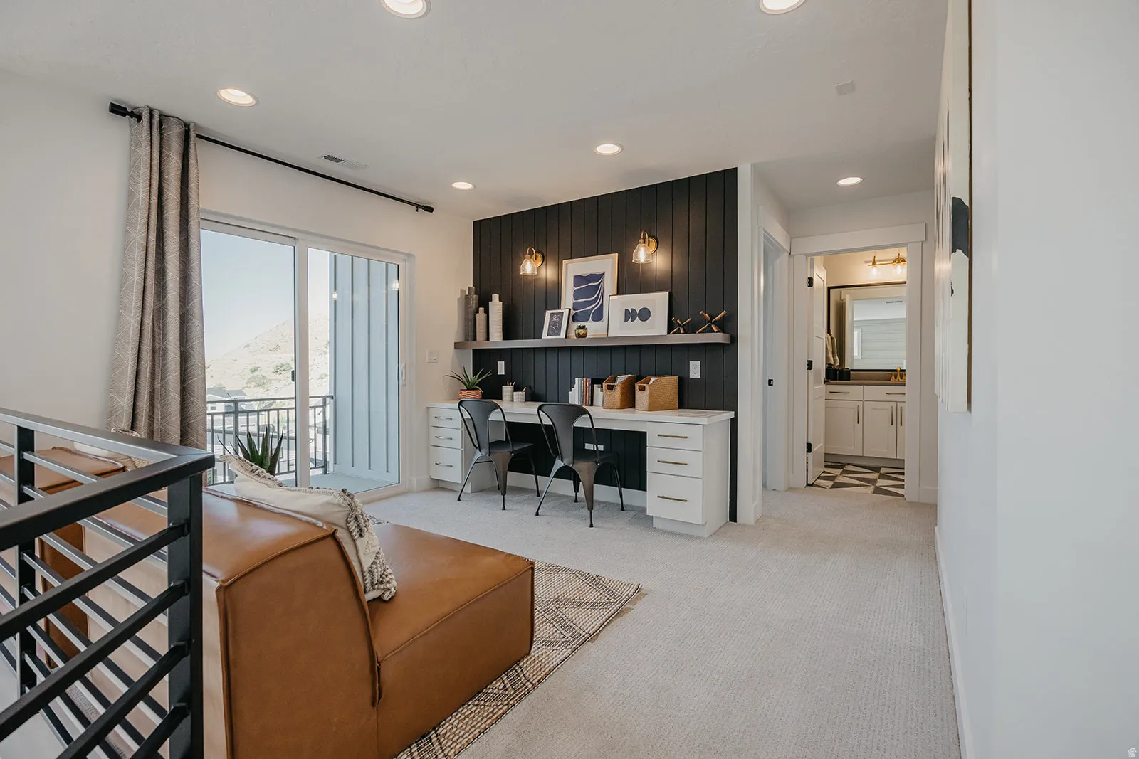 Upper-level living area with light-colored carpeting, featuring a built-in desk with shelving and sconce lighting against a dark accent wall