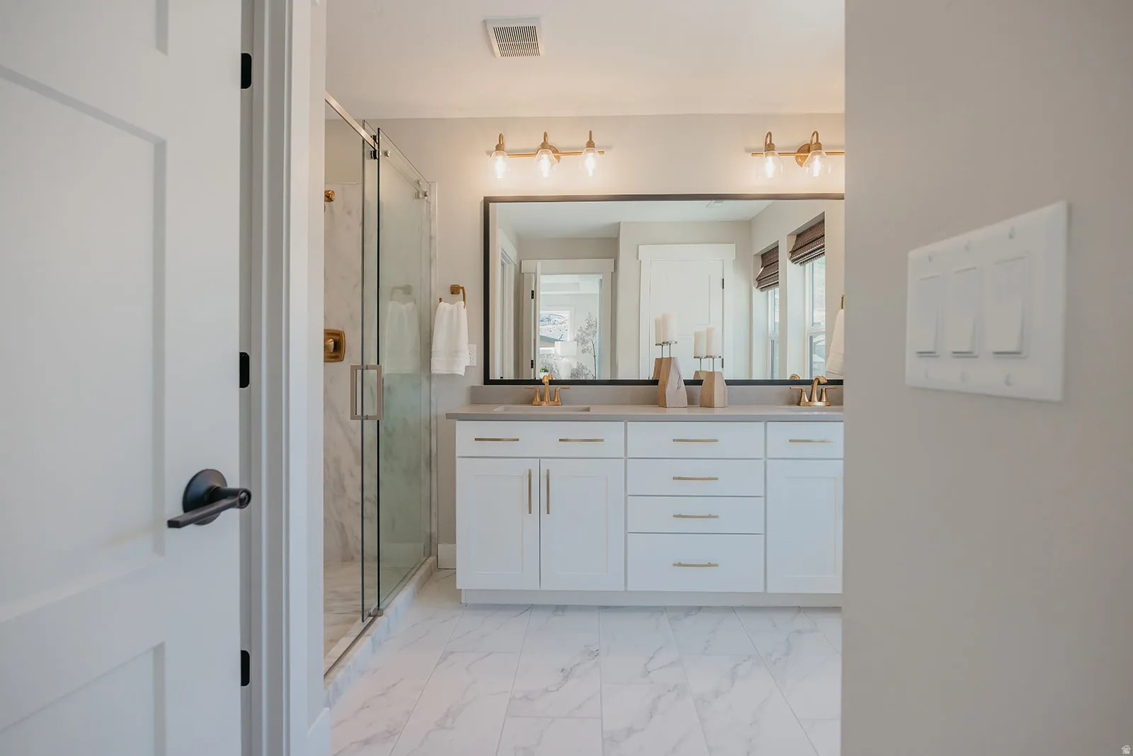 Bathroom featuring a dual vanity with white cabinetry and gold-finish hardware, a large framed mirror, a walk-in shower with a glass enclosure, and white marble-look tile flooring