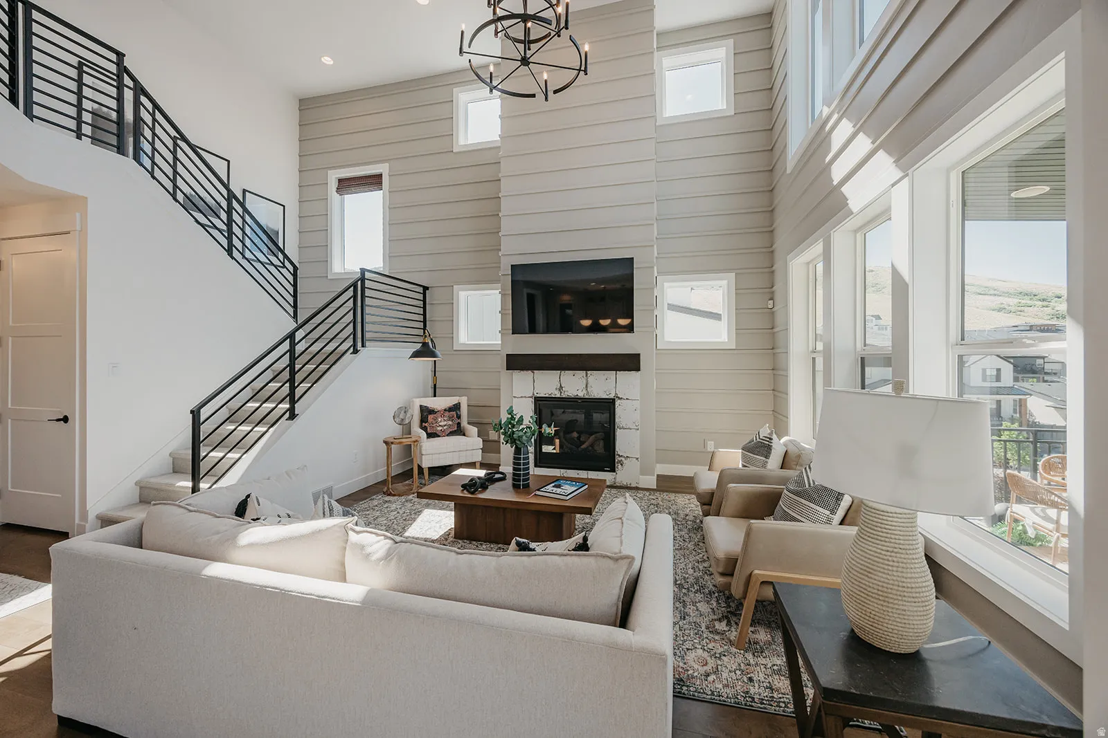 Two-story living area featuring a stone-clad fireplace, high ceilings, multiple large windows, a grand staircase with metal railings, and a contemporary chandelier