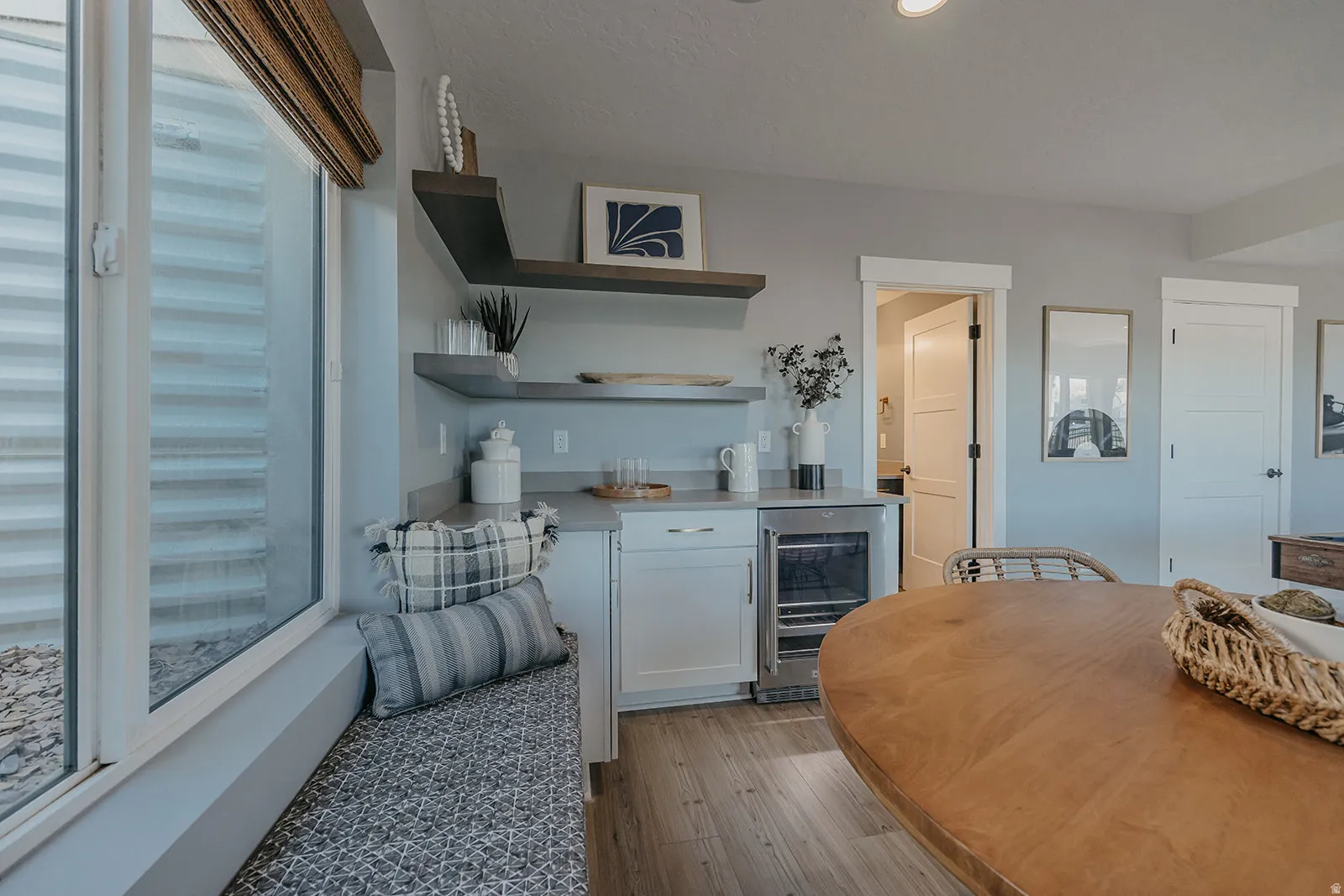 Built-in window bench with patterned upholstery, a wet bar featuring white cabinetry, a beverage refrigerator, and open shelving, all complemented by wood-finish flooring