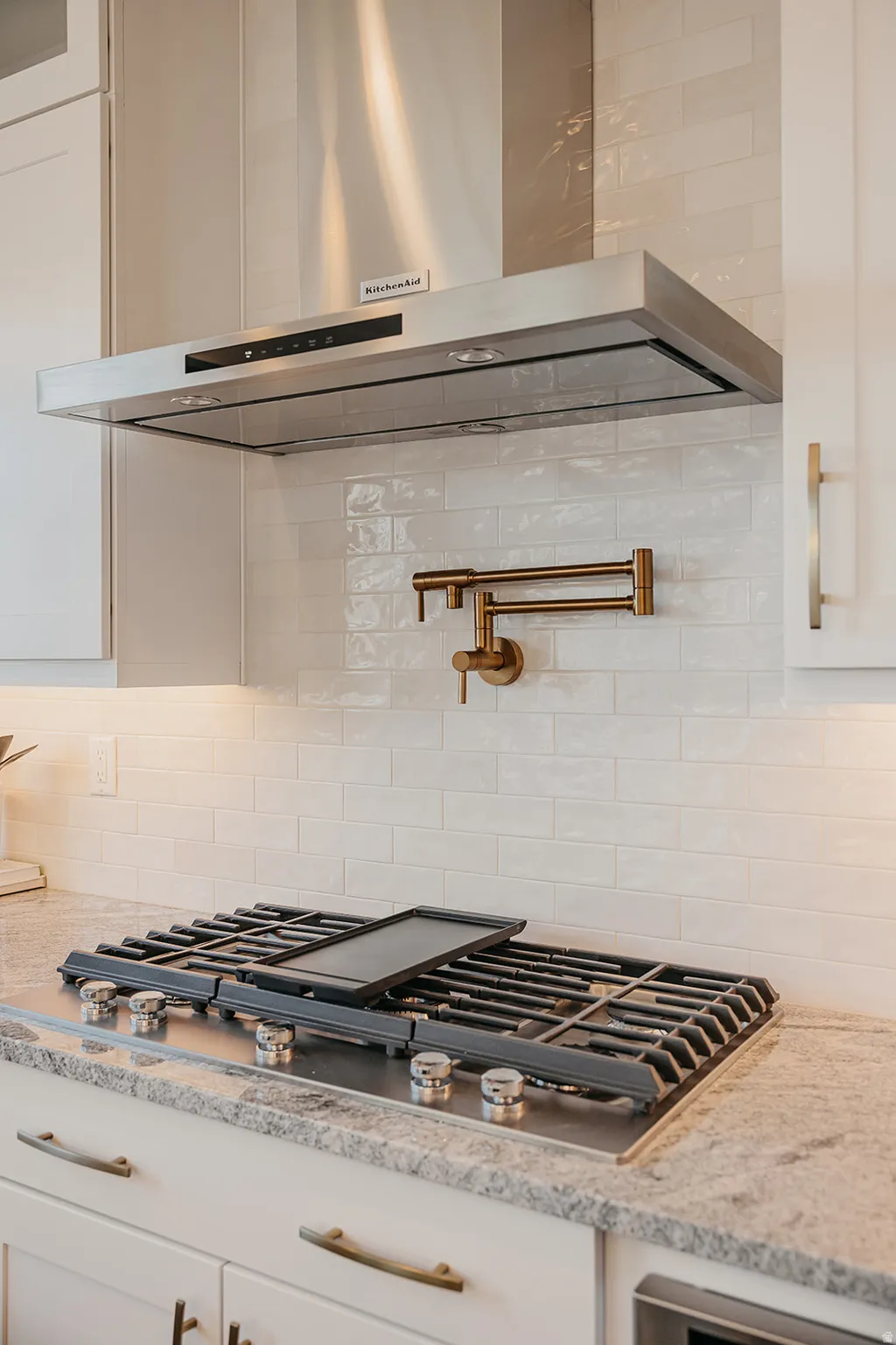 Stainless steel range hood above a gas cooktop with a griddle, complemented by a white subway tile backsplash and a bronze pot filler