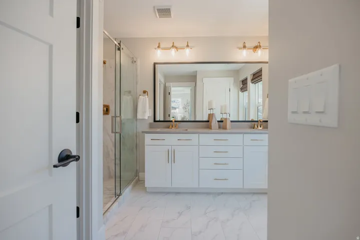 Bathroom featuring a dual vanity with white cabinetry and gold-finish hardware, a large framed mirror, a walk-in shower with a glass enclosure, and white marble-look tile flooring