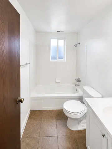 Unit A: Bathroom featuring a white bathtub, white tiled shower surround, frosted window, white vanity with integrated sink, and brown tile flooring