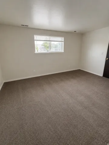 Unit A: Spacious room with neutral-toned carpet flooring, white baseboards, and a single window with blinds