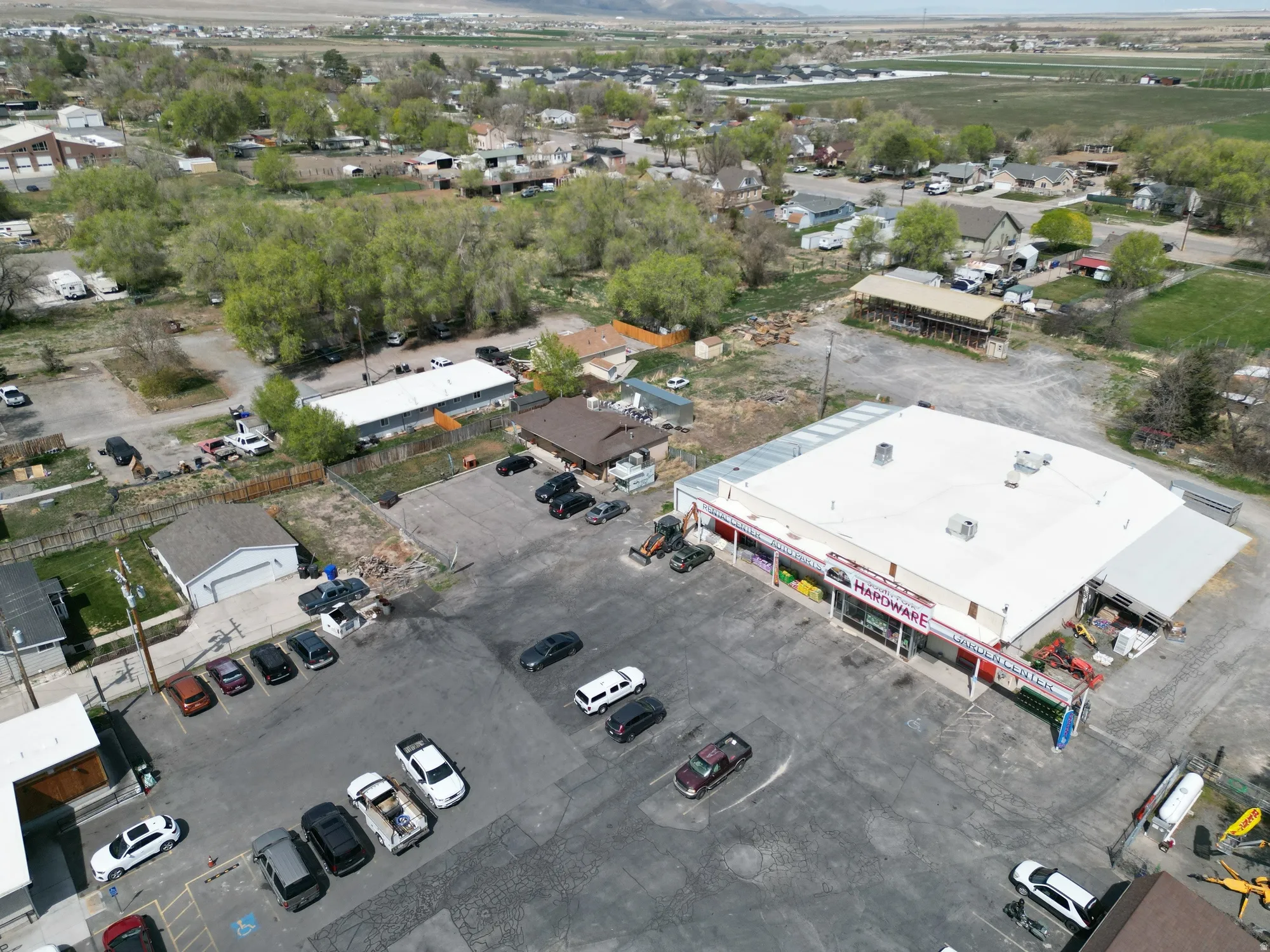 Commercial property featuring a large hardware store building, expansive asphalt parking lot, surrounding commercial structures, and a rural backdrop