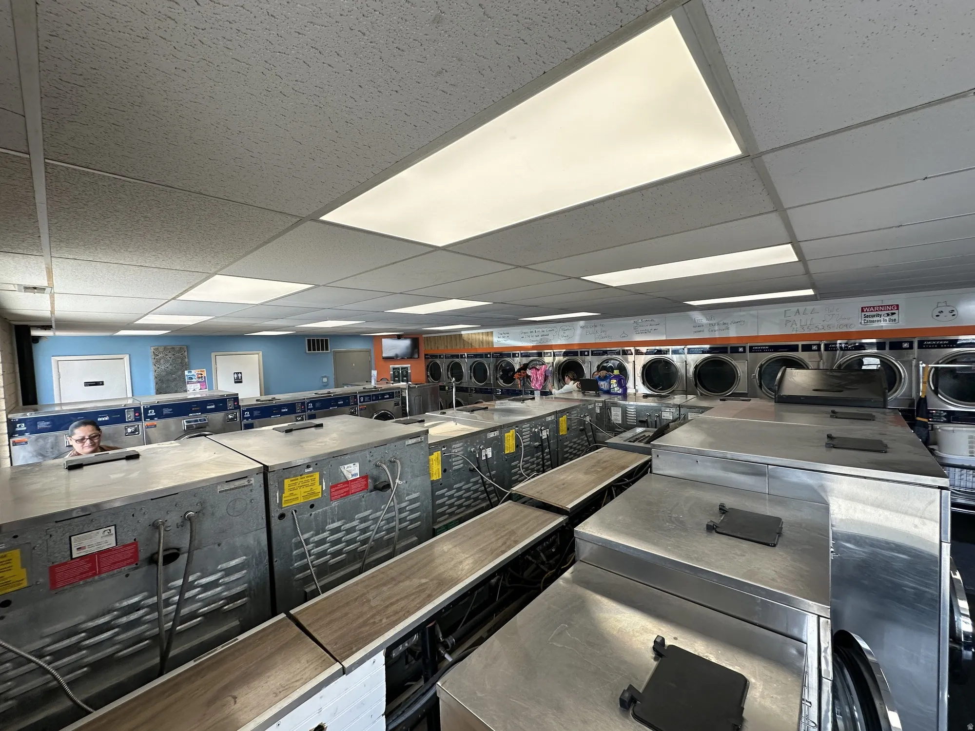 Commercial laundry facility featuring multiple front-load washing machines, fluorescent ceiling lighting, and a drop ceiling