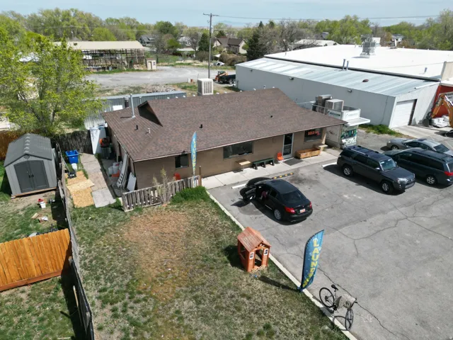 Single-story commercial building with a brown shingle roof and tan exterior, featuring an asphalt parking lot, fenced yard space, and an adjacent storage shed