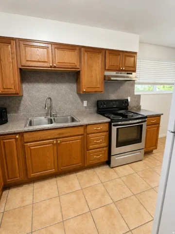 Unt A: Kitchen featuring wood-finish cabinetry with golden-toned hardware, a stainless steel double basin sink, a black electric range with a stainless steel oven, a coordinating stainless steel range hood, and tile flooring