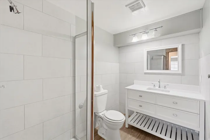 Modern bathroom featuring a glass-enclosed shower with white subway tile, a white vanity with integrated sink, a rectangular framed mirror, and wood-finish flooring