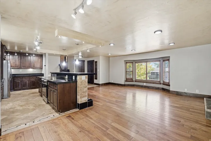 Spacious open-concept area featuring wood-finish flooring, a kitchen island with stone tile accents, stainless steel appliances, dark wood cabinetry, and a bay window with plantation shutters