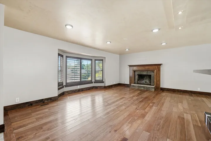 Expansive room featuring wide plank wood-finish flooring, a stone fireplace with a wood mantel, a bay window with plantation shutters, recessed lighting, and detailed baseboards