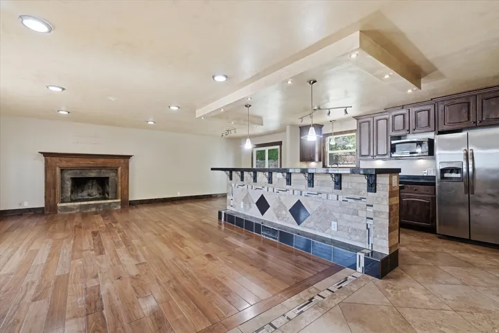 Open concept living space featuring wood-finish flooring, a stone fireplace with a wood mantle, a kitchen island with stone and tile accents, dark wood cabinetry, and stainless steel appliances