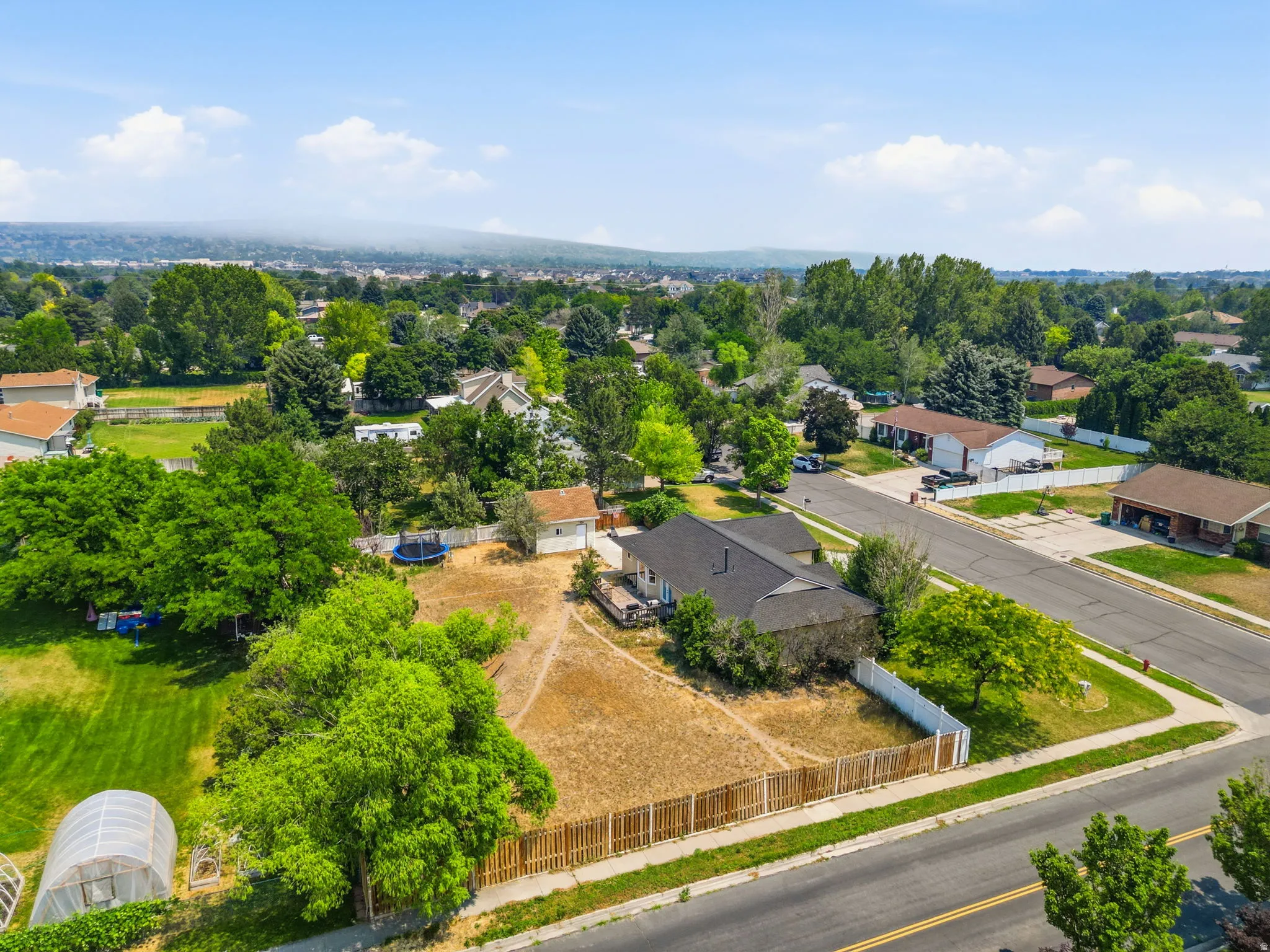 Expansive property featuring a large land parcel, a residential structure with a dark roof, a white storage shed, a greenhouse structure, and mature trees