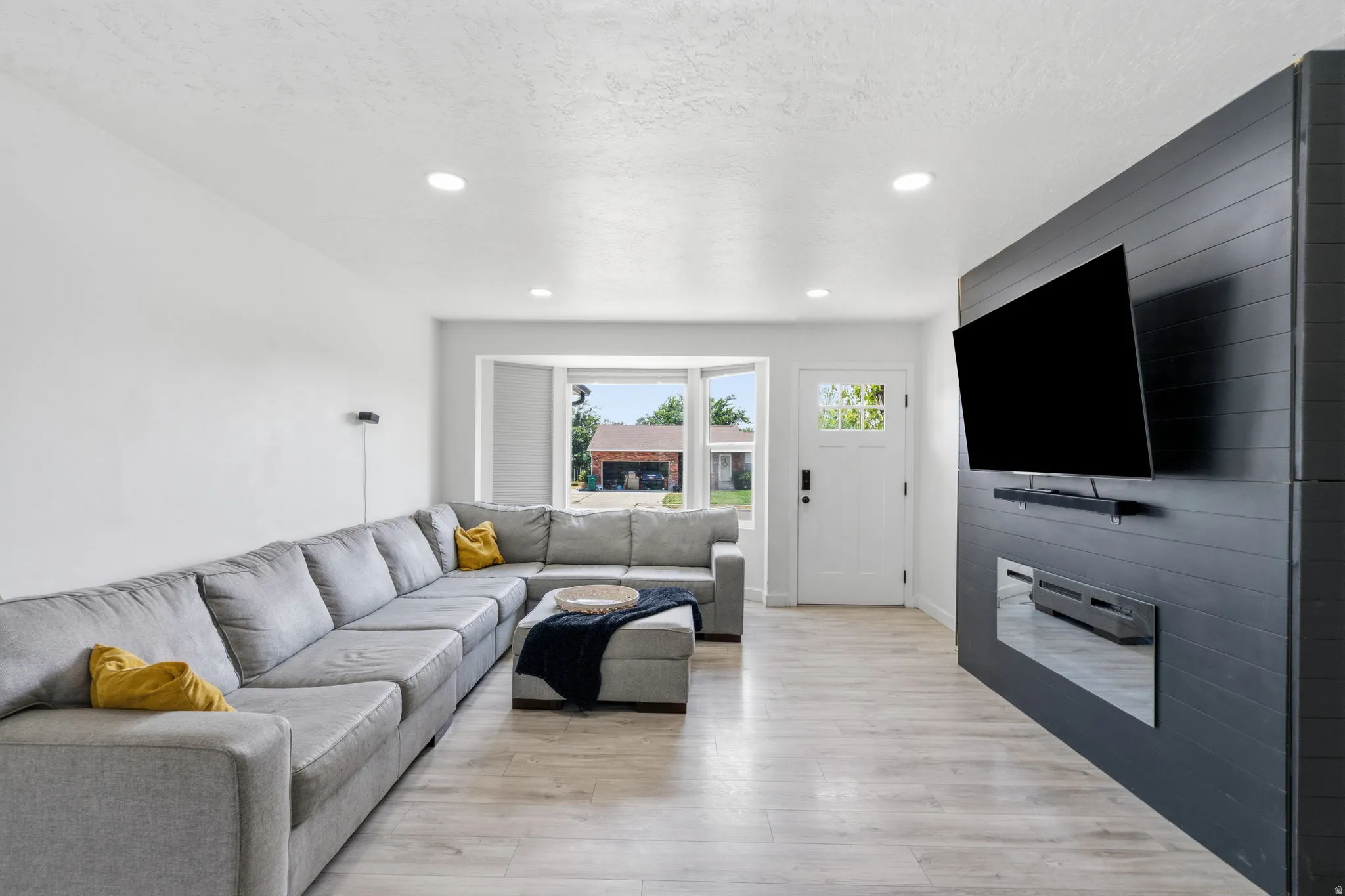 Spacious living area featuring light wood-finish flooring, recessed lighting, a modern built-in entertainment wall with an integrated electric fireplace, and a bay window