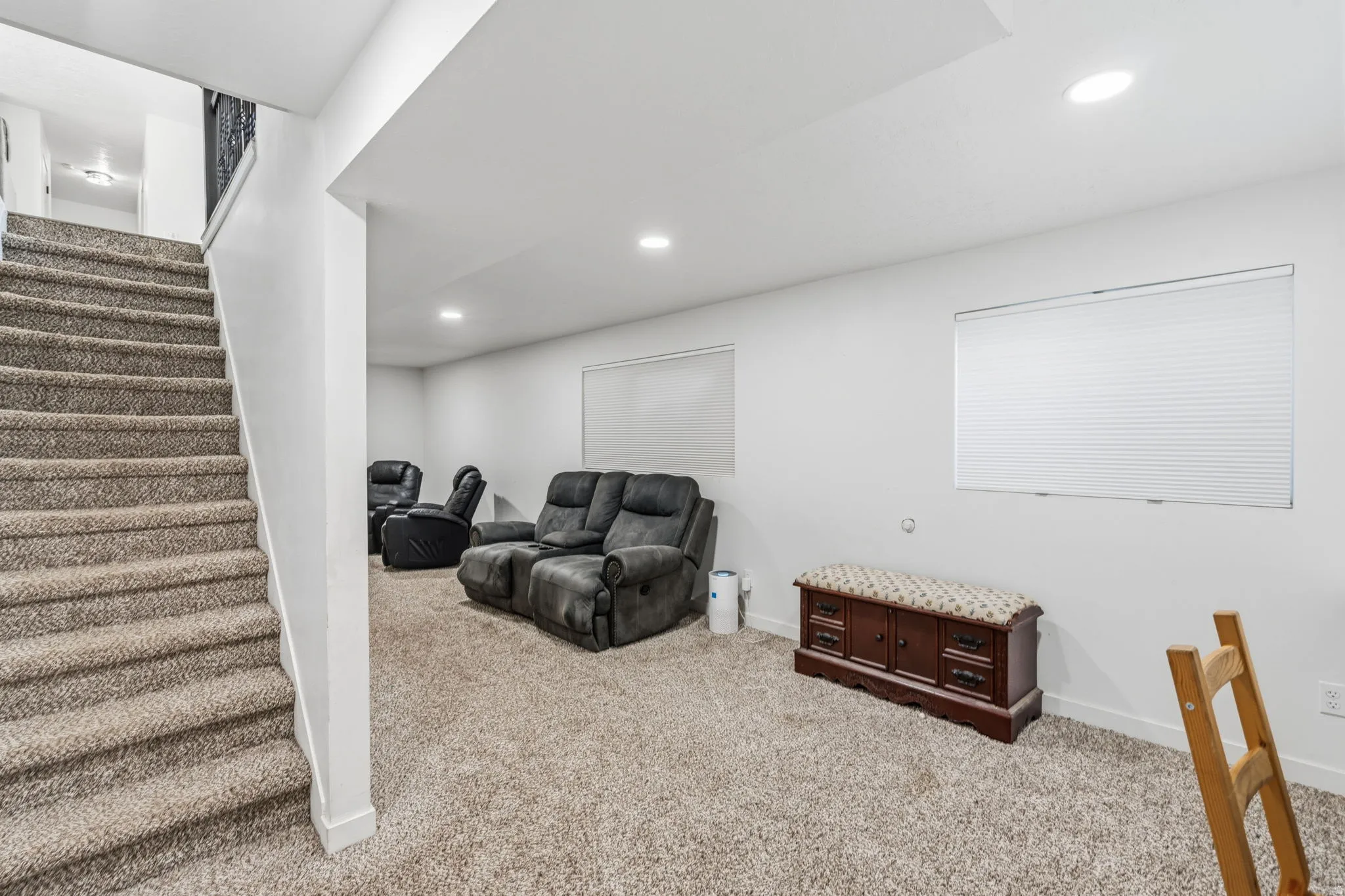 Carpeted staircase descending into a spacious lower-level room featuring recessed lighting, two windows with cellular blinds, and neutral wall finishes