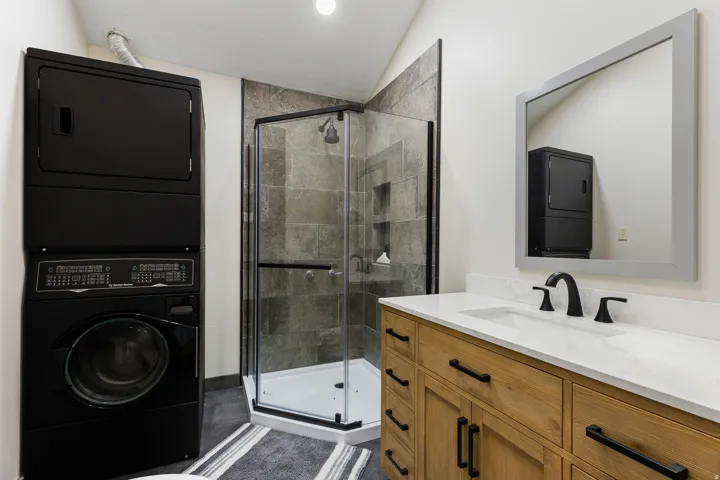 Bathroom featuring a stacked washer and dryer, corner shower with dark-framed glass enclosure, and a wood-finish vanity with a white countertop and matte black fixtures