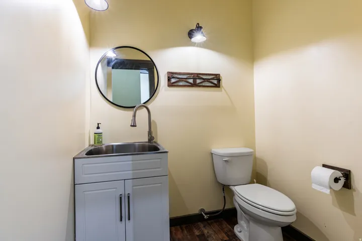 Compact powder room featuring wood-finish flooring, a white vanity with stainless steel sink, a circular mirror, and a wall-mounted toilet