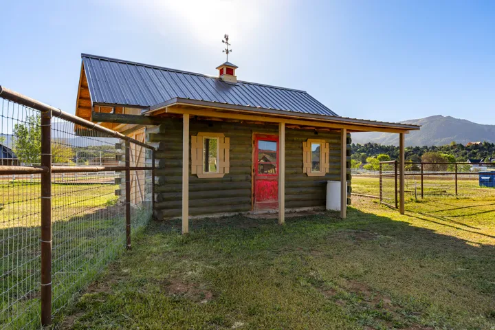 Log cabin style outbuilding featuring a red door, light wood window shutters, a covered porch, and a metal roof with a decorative weathervane