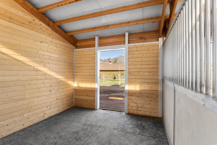 Interior stable space featuring light wood paneling, exposed wood ceiling beams, a concrete floor, and a metal-barred partition