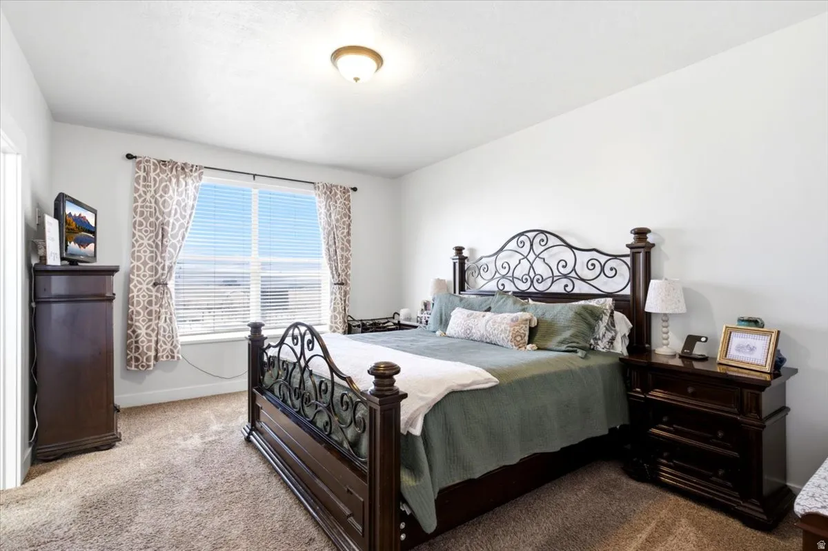 Carpeted bedroom featuring a large window with blinds, ceiling-mounted light fixture, and white wall finishes