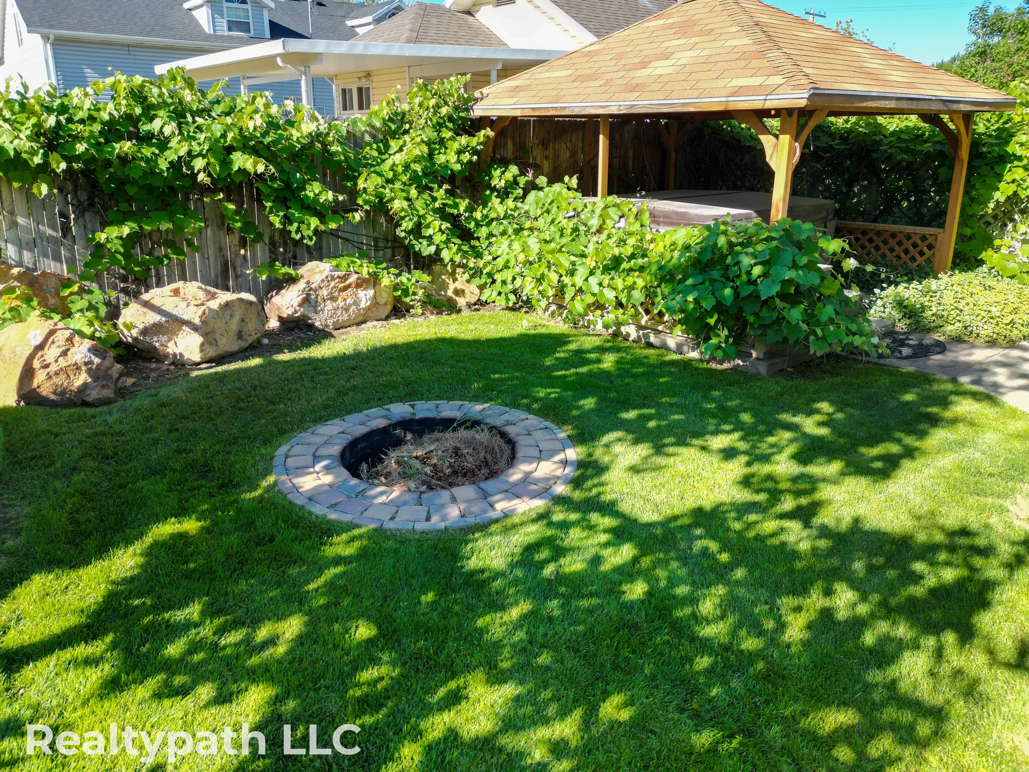 Manicured backyard featuring a stone fire pit, a wooden gazebo with a shingle roof, and a privacy fence lined with mature foliage