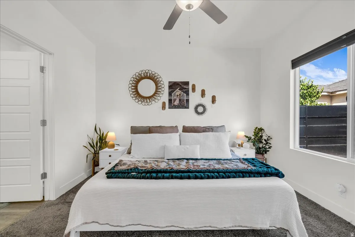 Bright bedroom featuring a ceiling fan, modern window with external fencing, white paneled door, and neutral-toned carpeting