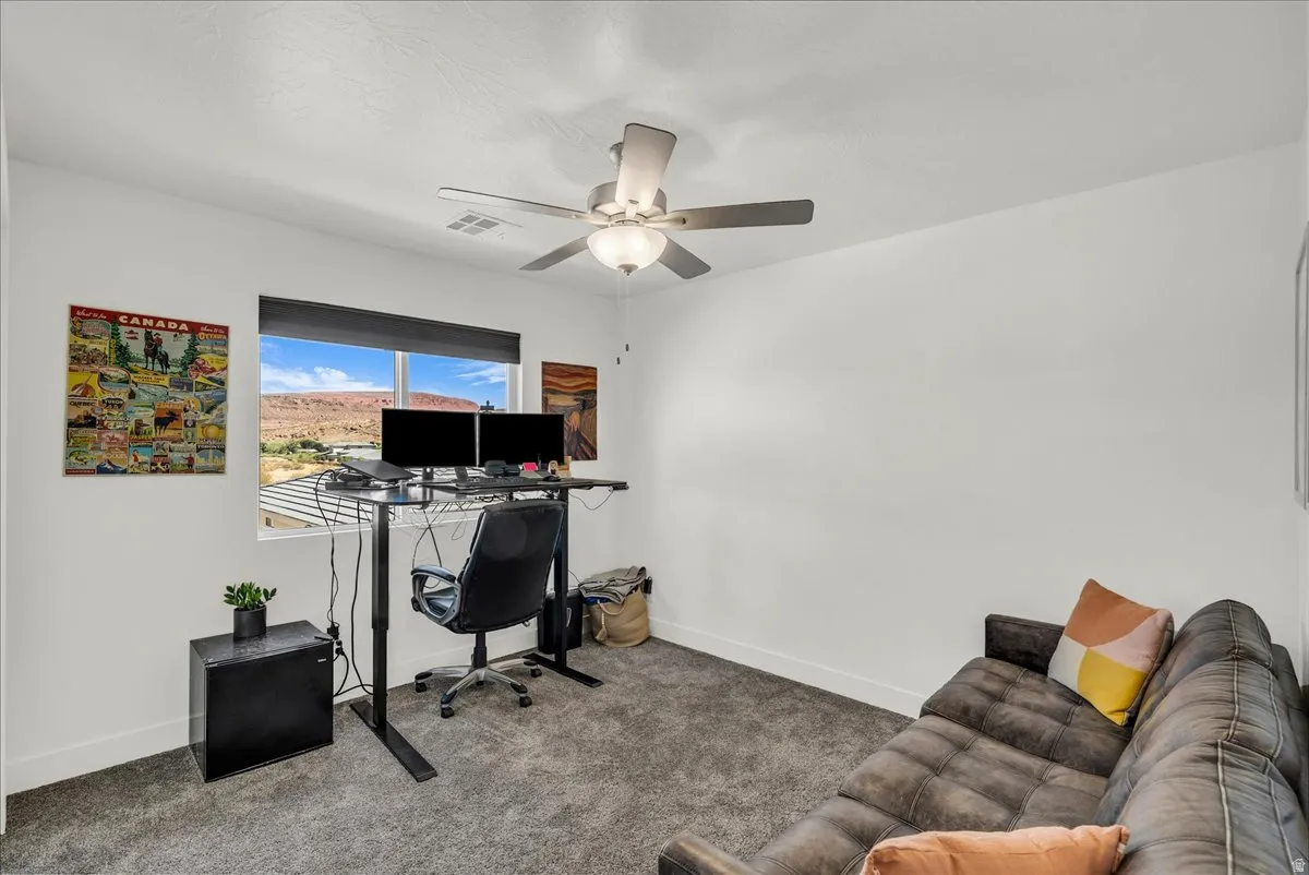 Interior space featuring a window with expansive distant geological formations, ceiling fan with light fixture, white walls, dark gray carpet, and white baseboards