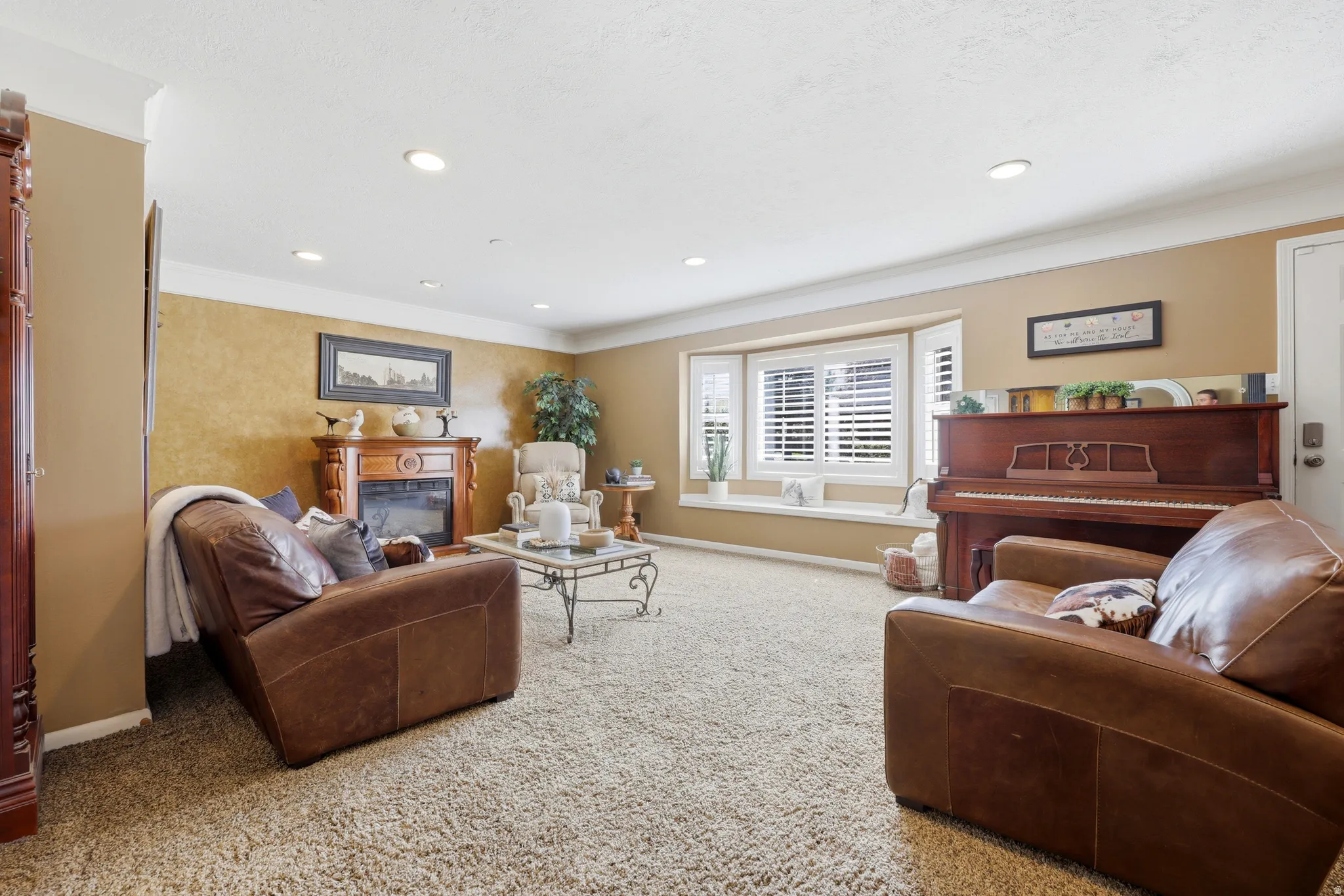Spacious living area featuring a fireplace with a wood mantel, a bay window with plantation shutters, recessed lighting, and crown molding