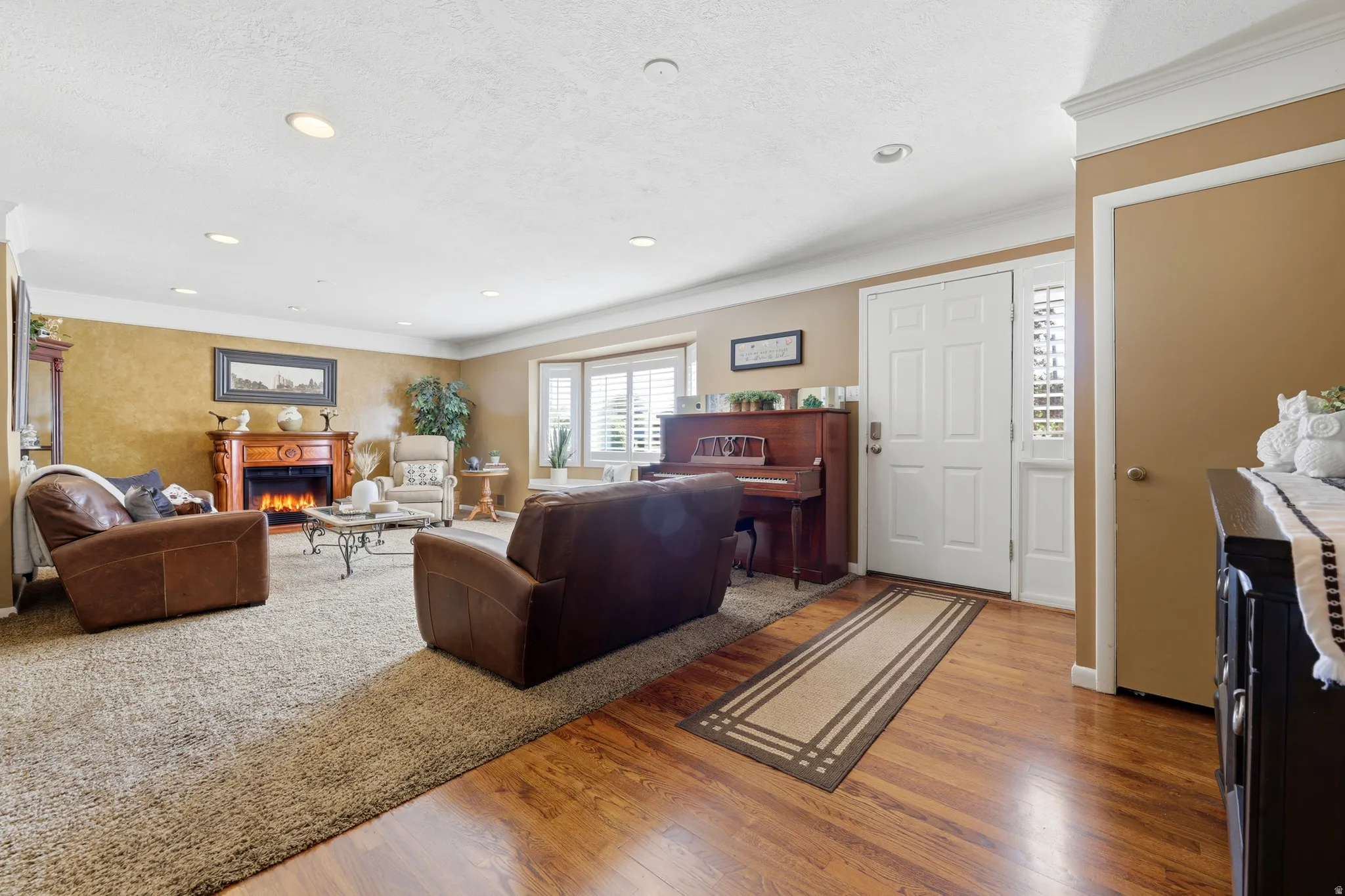 Warm living space featuring a decorative fireplace with wood mantel, a bay window with plantation shutters, recessed lighting, and a white paneled entry door with sidelight