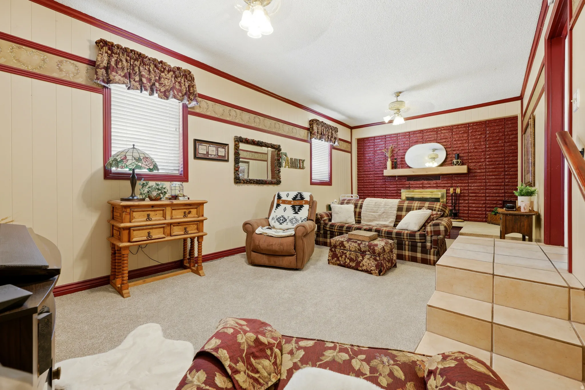 Living area featuring a brick accent wall with fireplace, neutral carpet flooring, a drop ceiling with a ceiling fan, and tiled steps