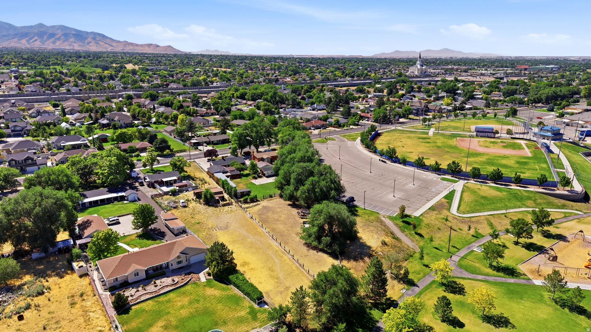 Expansive residential area featuring a baseball field, open-air park, paved walking paths, a large parking area, and distant mountain ranges