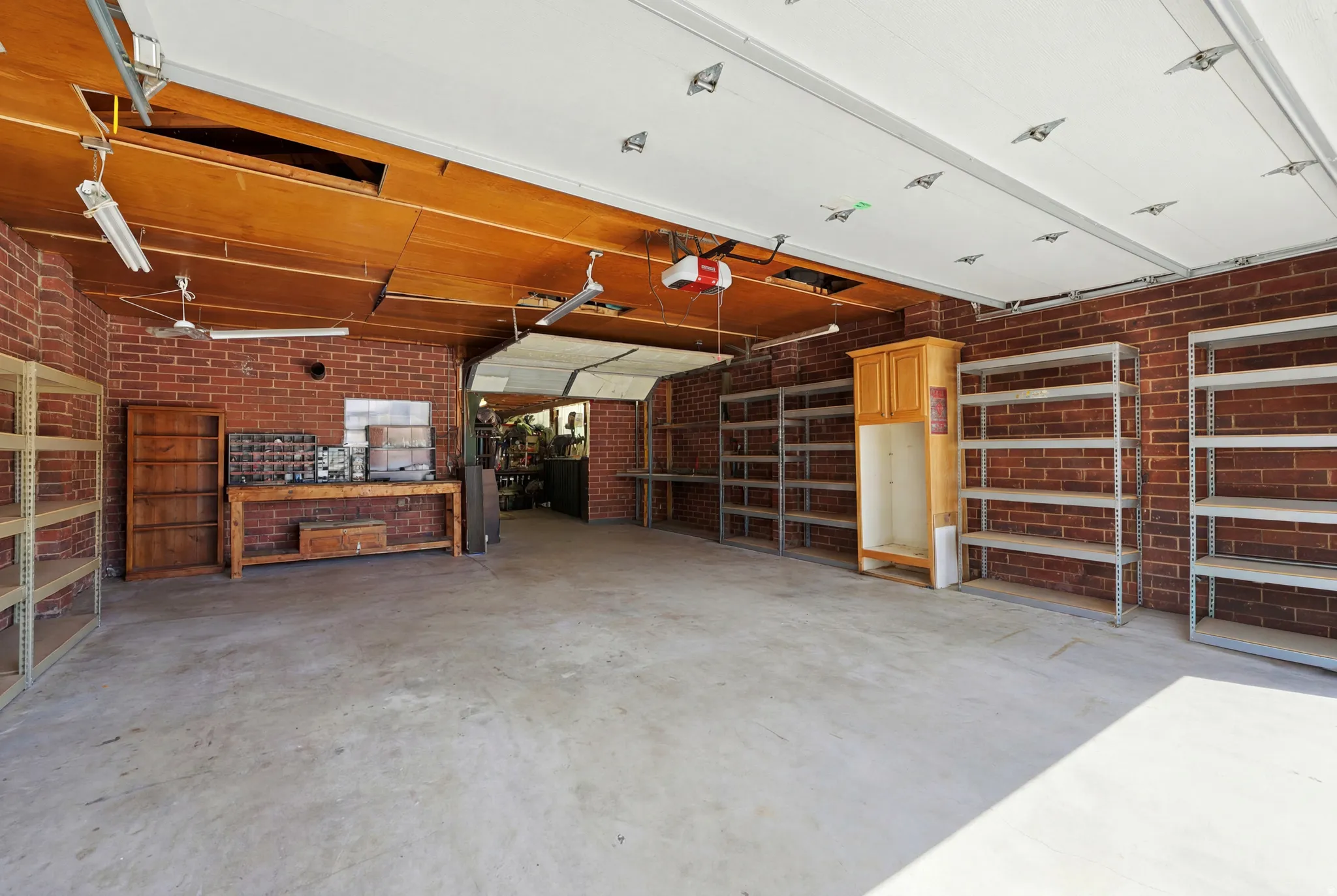 Spacious garage featuring brick walls, concrete flooring, extensive shelving, overhead lighting, and an automatic garage door opener