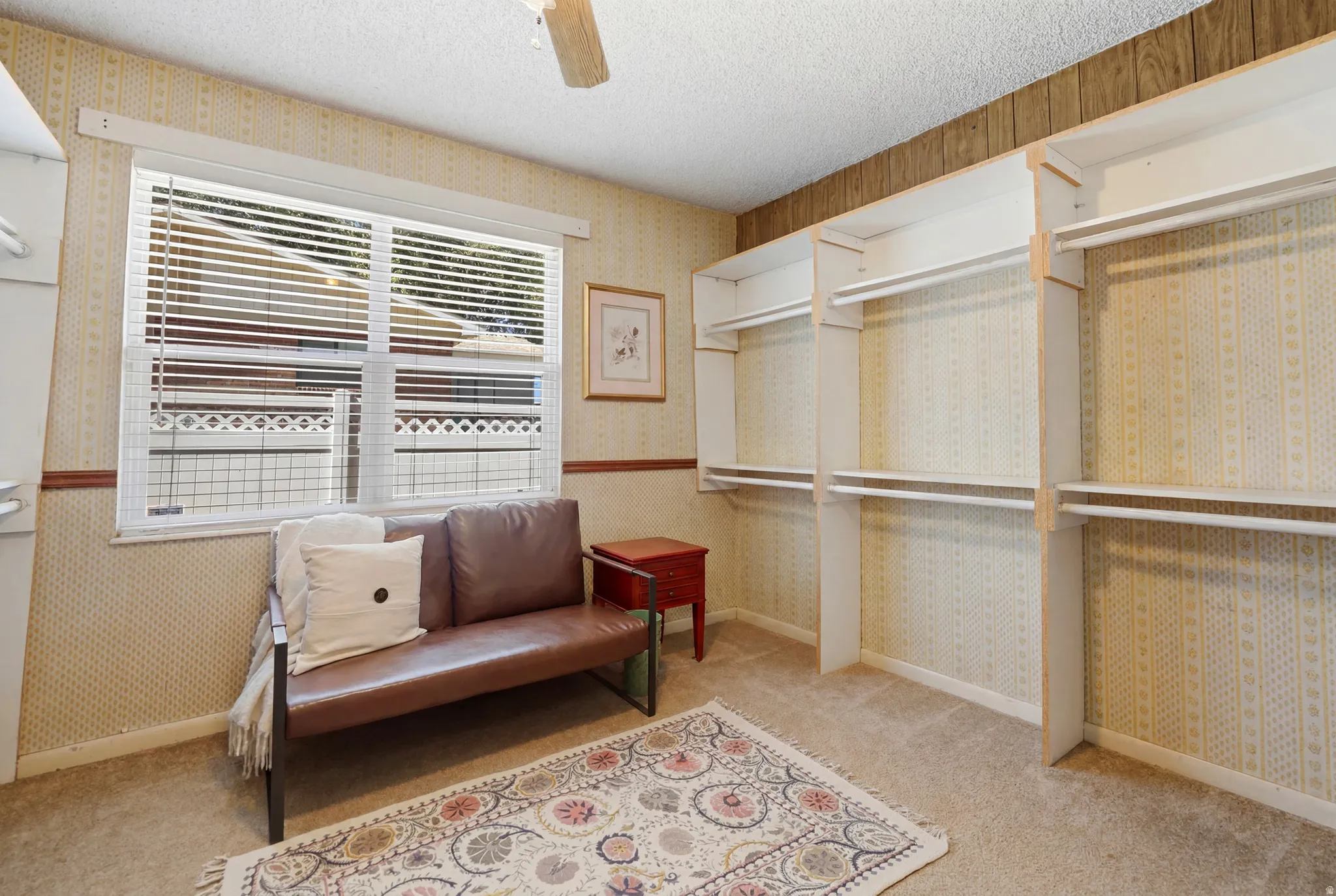 Carpeted room featuring a large window with blinds, extensive built-in shelving, and a ceiling fan