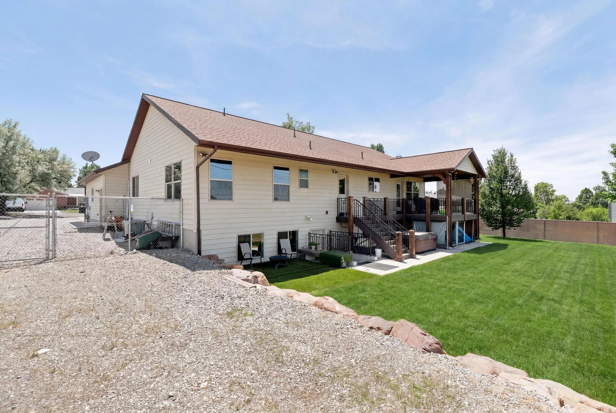Expansive backyard featuring a manicured lawn, a multi-level deck with a covered portion, and a gravel driveway with chain-link fencing