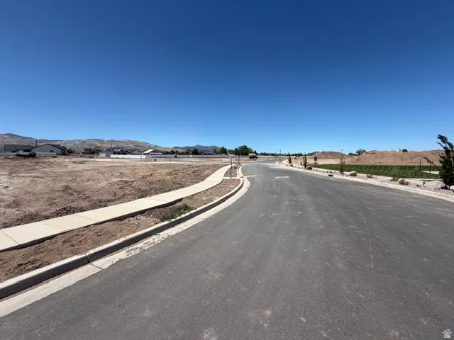 Paved street with concrete curbing and sidewalks, bordered by undeveloped land and distant mountains
