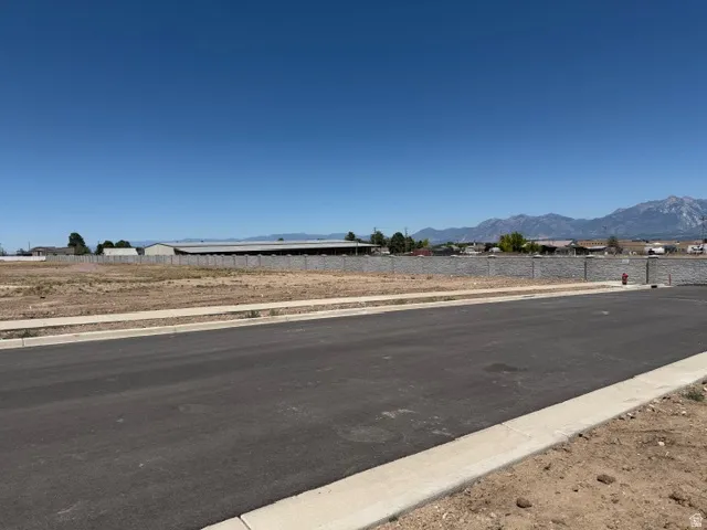Expansive undeveloped lot featuring a paved road, concrete sidewalk, low retaining wall, and mountain backdrop