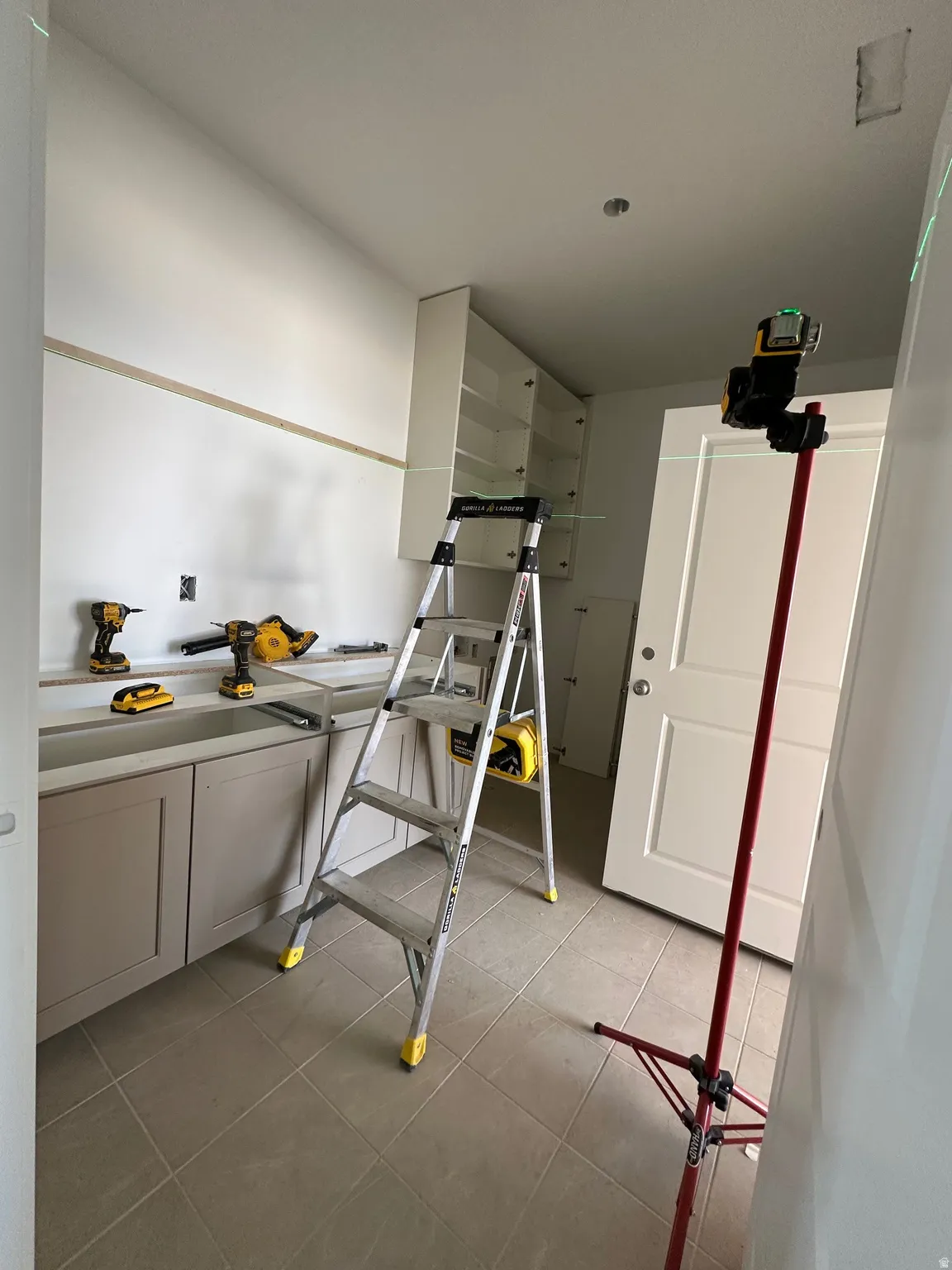 Unfinished utility room featuring light-toned floor tiling, lower cabinetry with shaker-style doors, and upper wall-mounted shelving