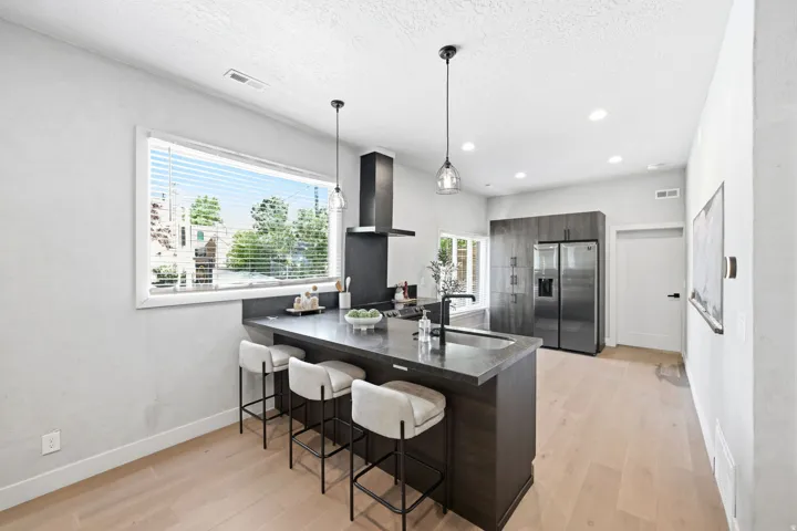 Modern kitchen featuring a large central island with integrated sink, black range hood, stainless steel refrigerator, dark wood-finish cabinetry, and light wood-finish flooring