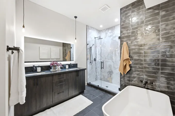 Contemporary bathroom featuring a freestanding soaking tub, a frameless glass shower enclosure with marble-look tile, a dual-sink vanity with dark wood-finish cabinetry and a stone countertop, and an illuminated mirror